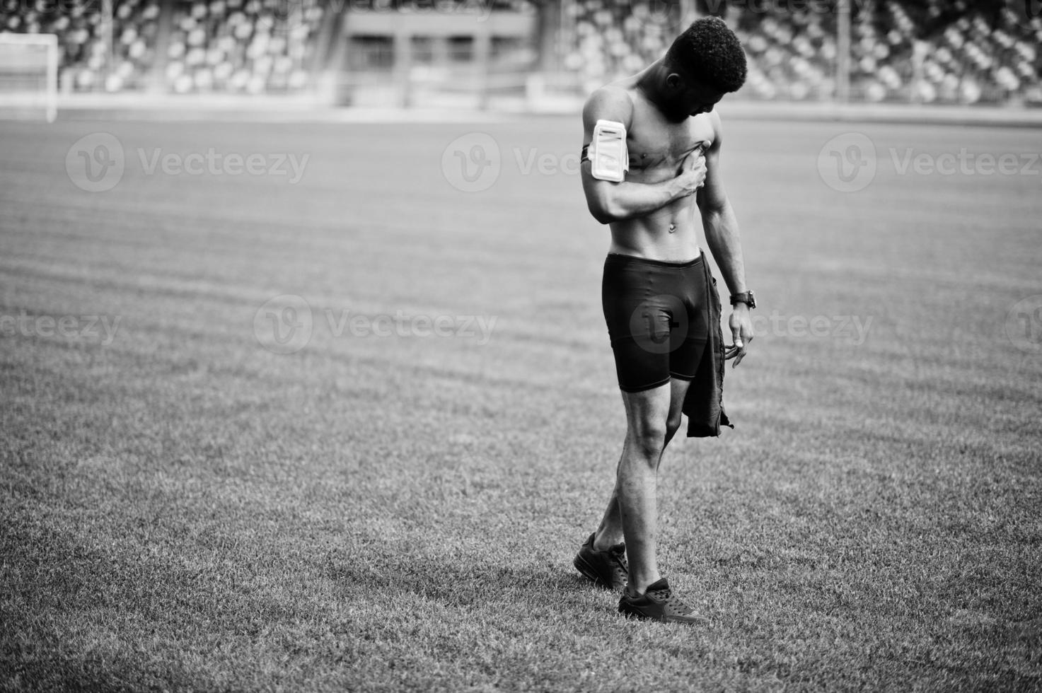 African american male athlete sexy sport bare torso man with running sports arm case for mobile phone, posed at green grass of football stadium. photo