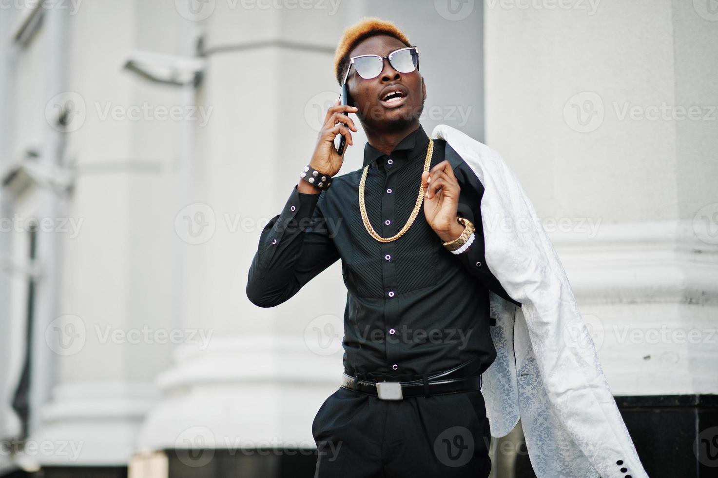 hombre afroamericano elegante y guapo con traje blanco y camisa negra con teléfono móvil. foto