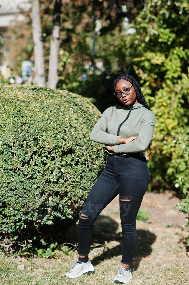 City portrait of positive young dark skinned female wearing green hoody and eyeglasses against greenery. photo