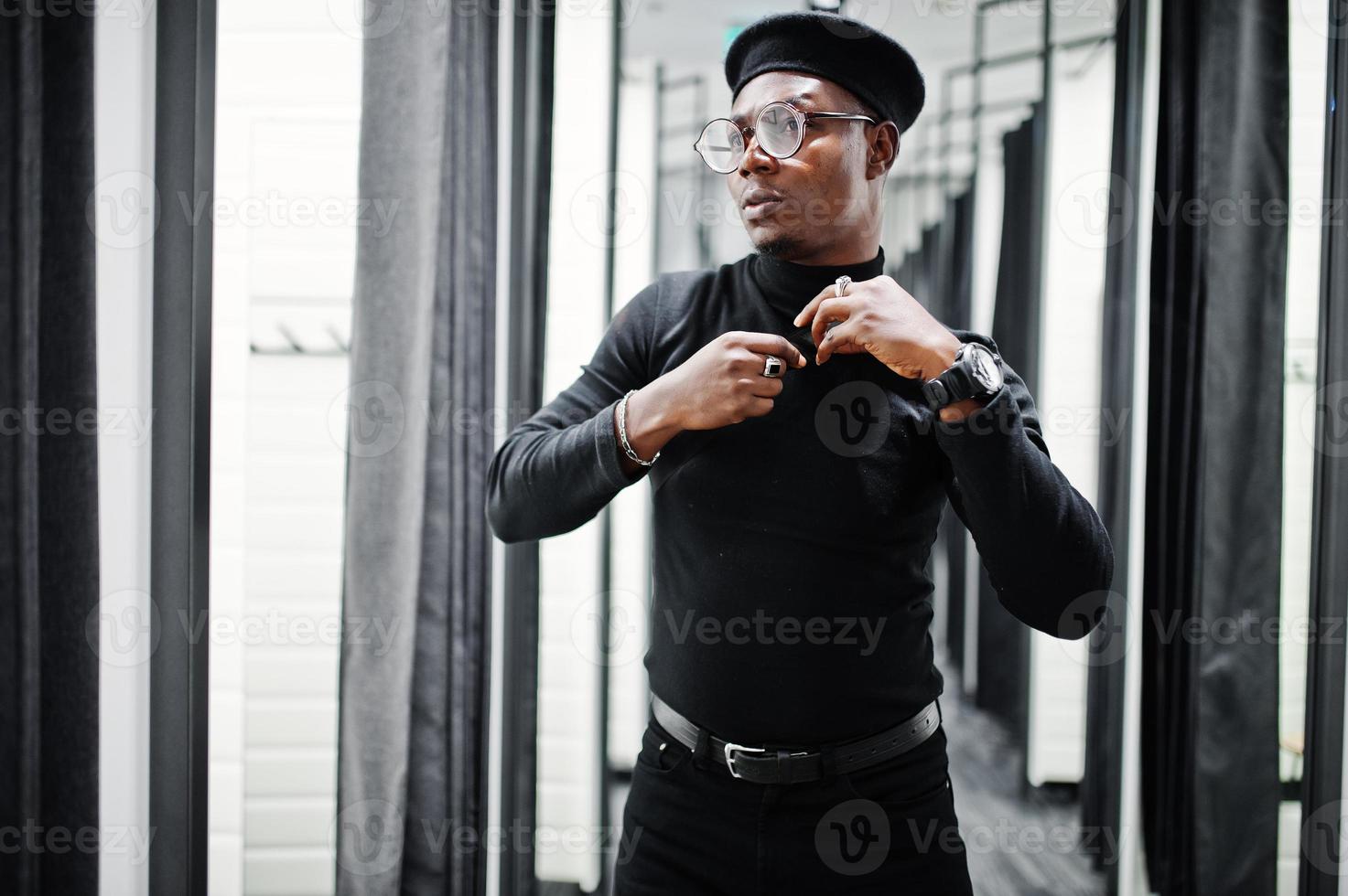 Elegante hombre afroamericano casual con traje negro y boina con riñonera en la tienda de ropa de la sala de montaje, mirando al espejo. foto