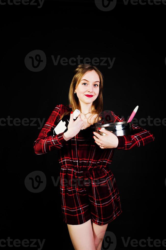 Young housewife in checkered dress with saucepan and kitchen spoon and bottle isolated on black background. photo