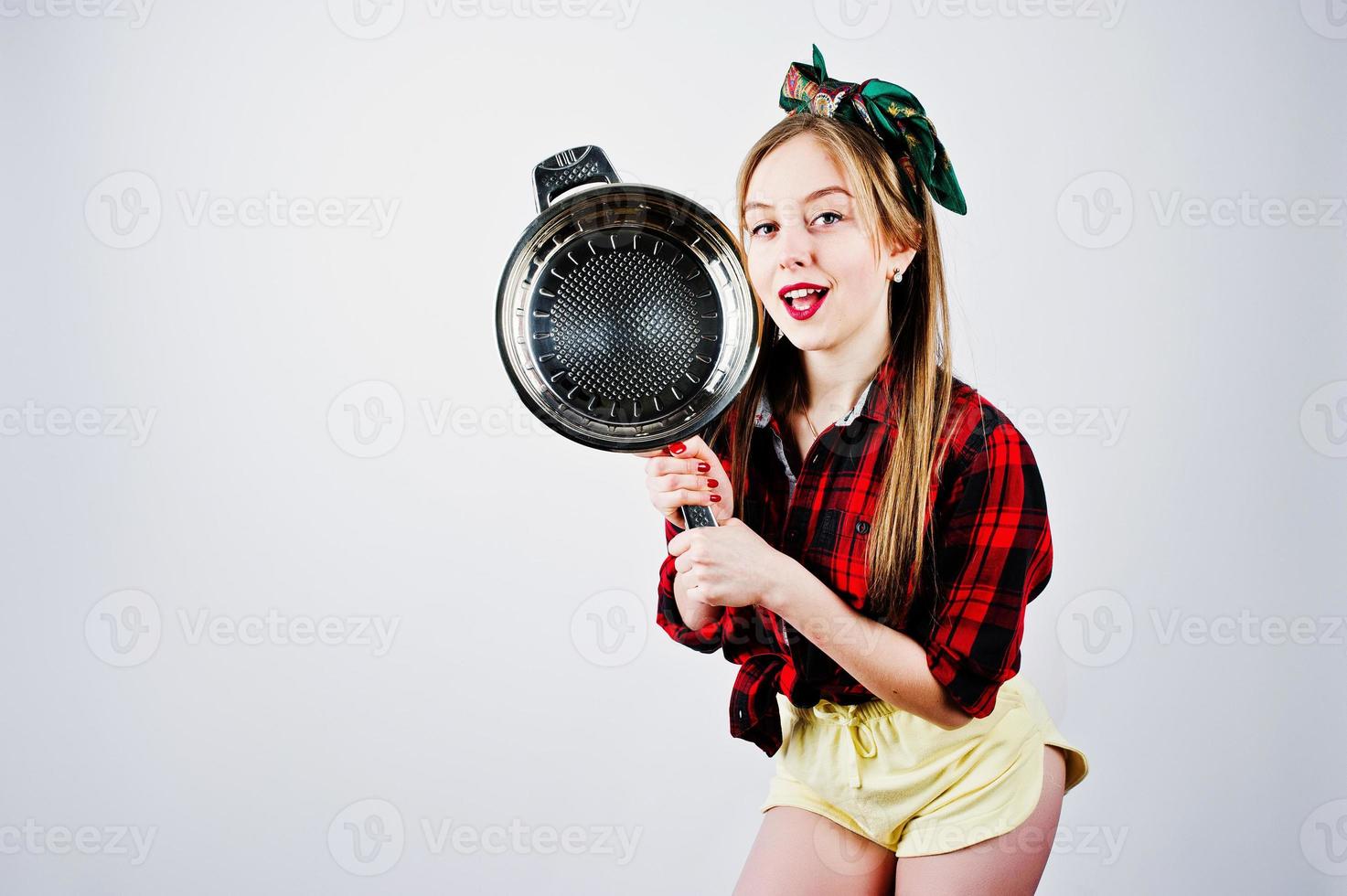 joven ama de casa divertida en camisa a cuadros y pantalones cortos amarillos pin up estilo con sartén aislado sobre fondo blanco. foto