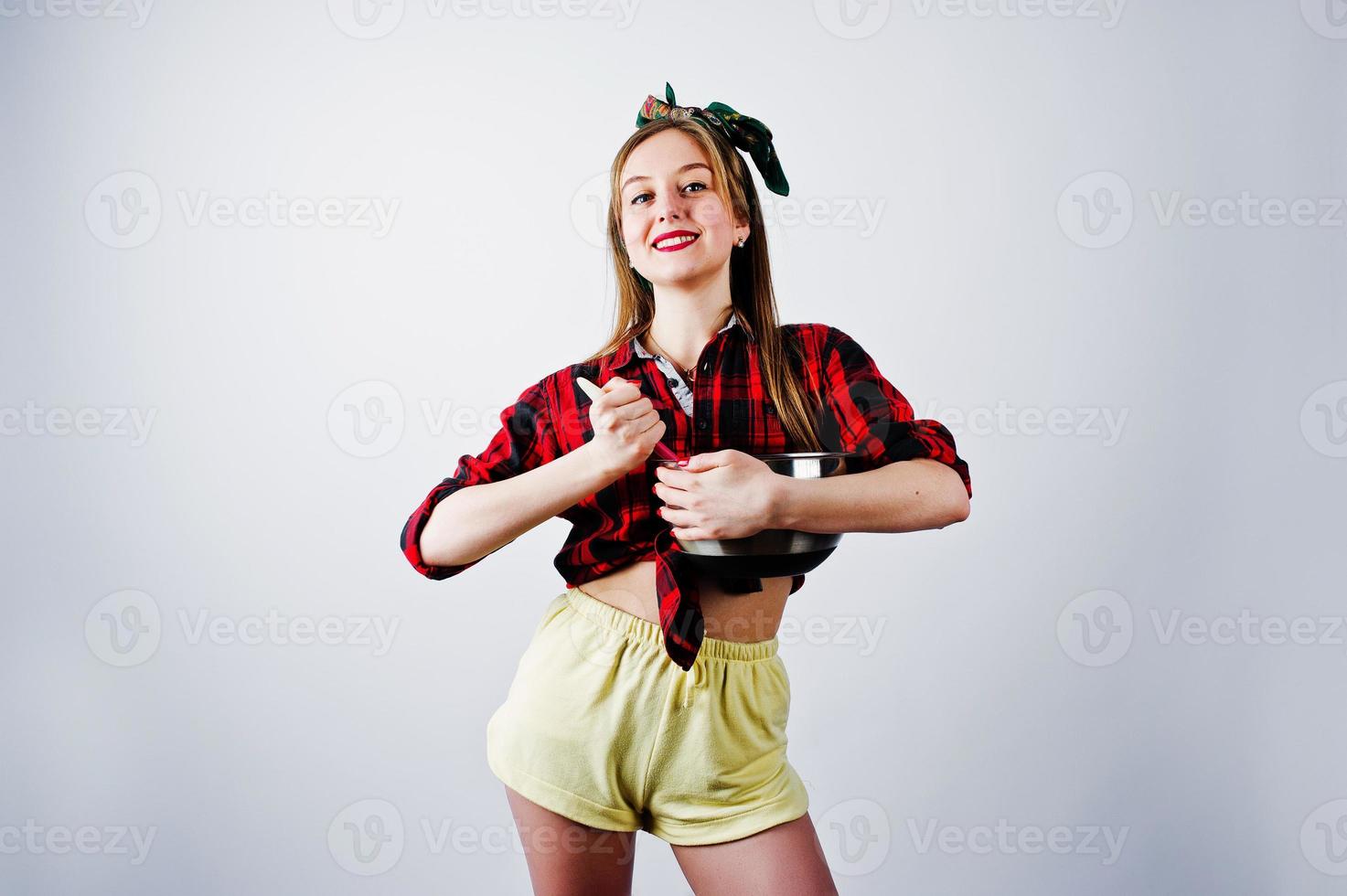 joven ama de casa divertida en camisa a cuadros y pantalones cortos amarillos pin up estilo con cacerola y cuchara de cocina aislado sobre fondo blanco. foto