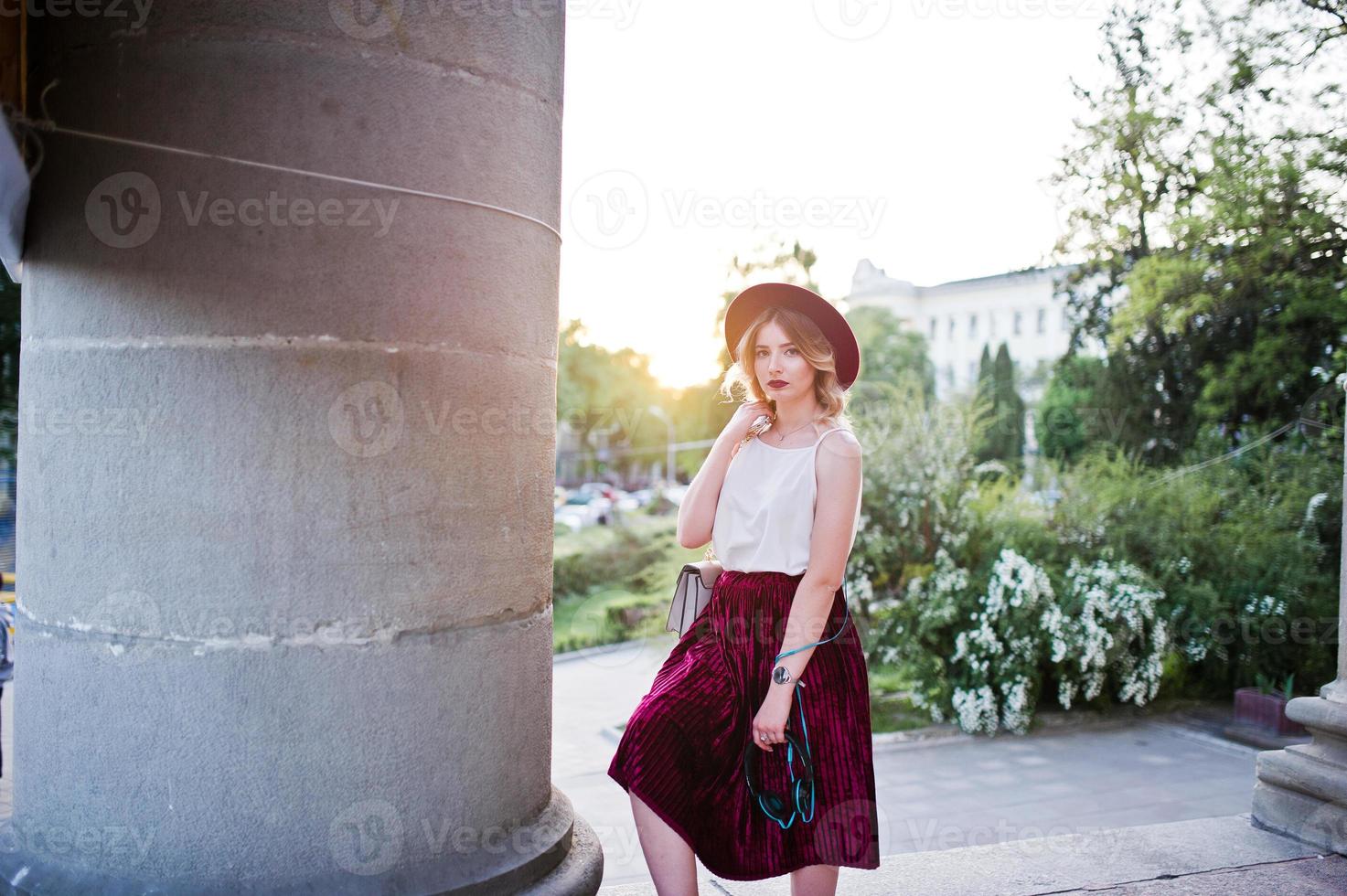 Fashionable and beautiful blonde model girl in stylish red velvet velour skirt, white blouse and hat, posed with phone and earphones against column at sunset. photo