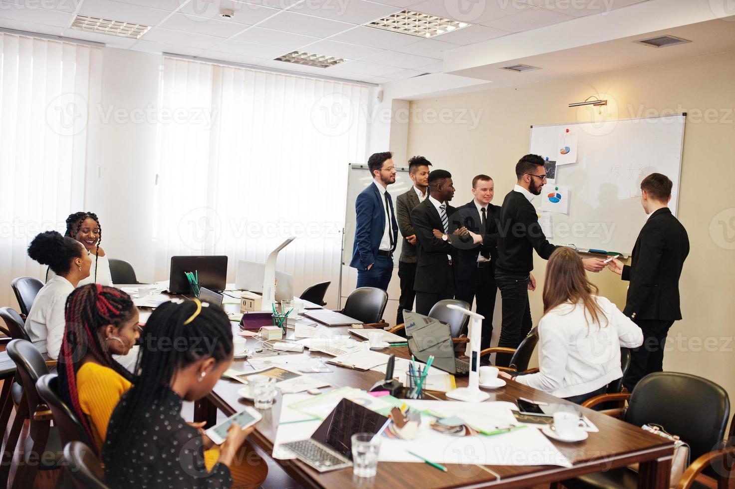 Multiracial business team addressing meeting around boardroom table and write plan on board. photo