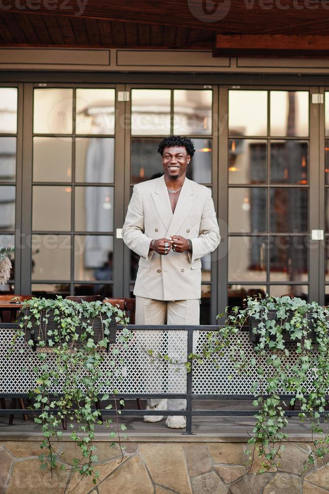 Stylish afro man in beige old school suit standing on terrace of cafe. Fashionable young African male in casual jacket on bare torso. photo