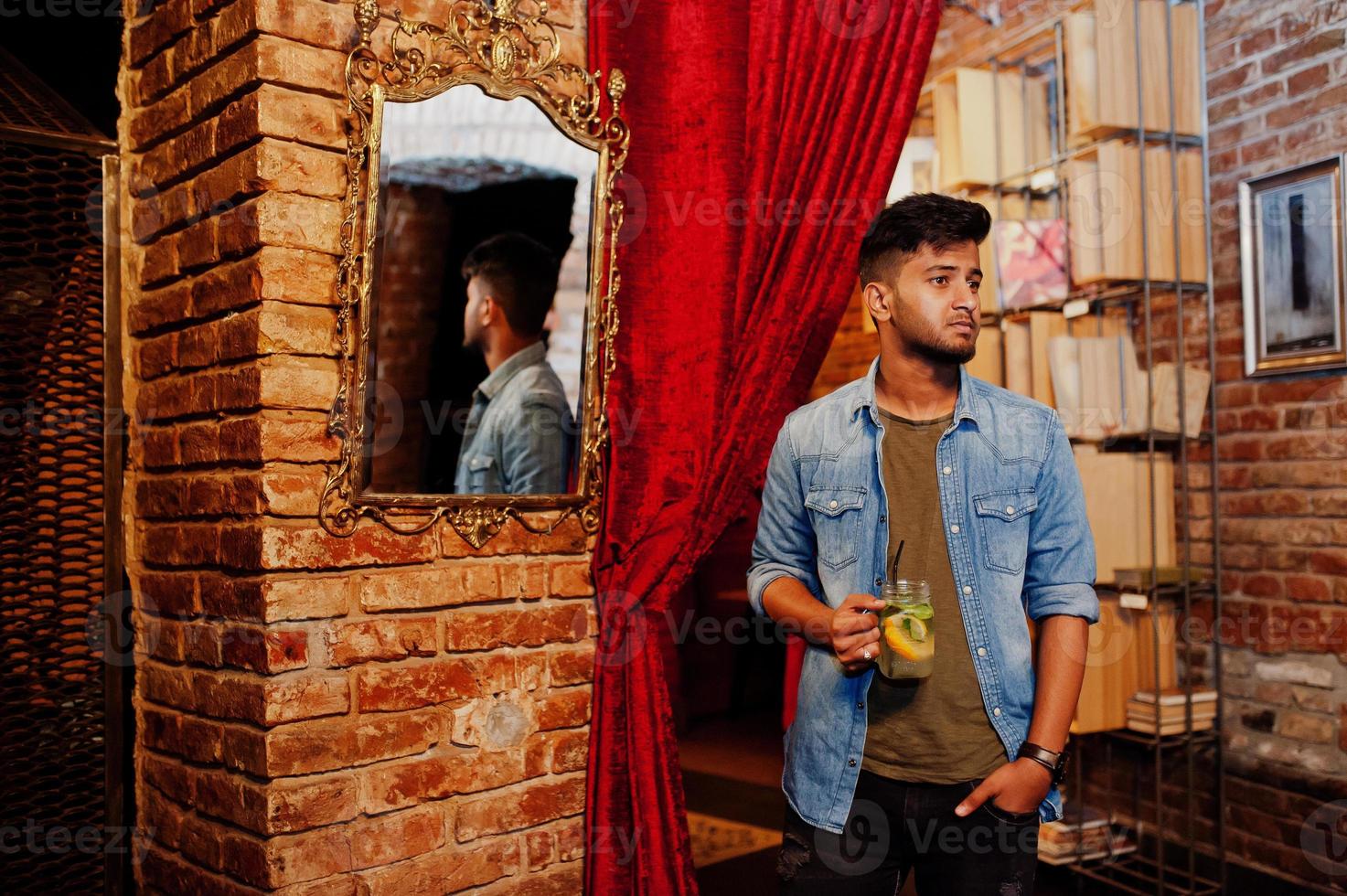 Portrait of handsome successful bearded south asian, young indian freelancer in blue jeans shirt standing in cafe with cocktail against mirror. photo