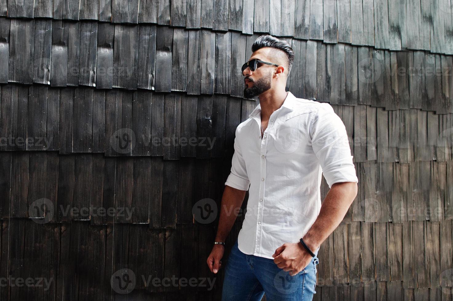 Stylish tall arabian man model in white shirt, jeans and sunglasses posed against wooden wall indoor. Beard attractive arab guy. photo