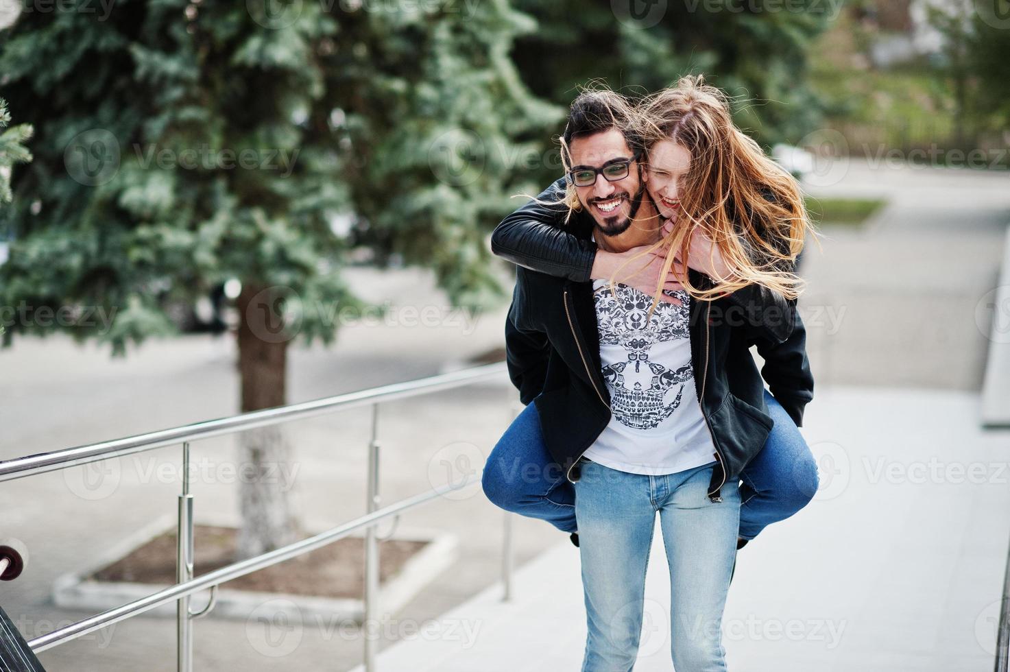 Cool multiracial couple posing with longboard. European girl jumped over shoulder arabian man. photo