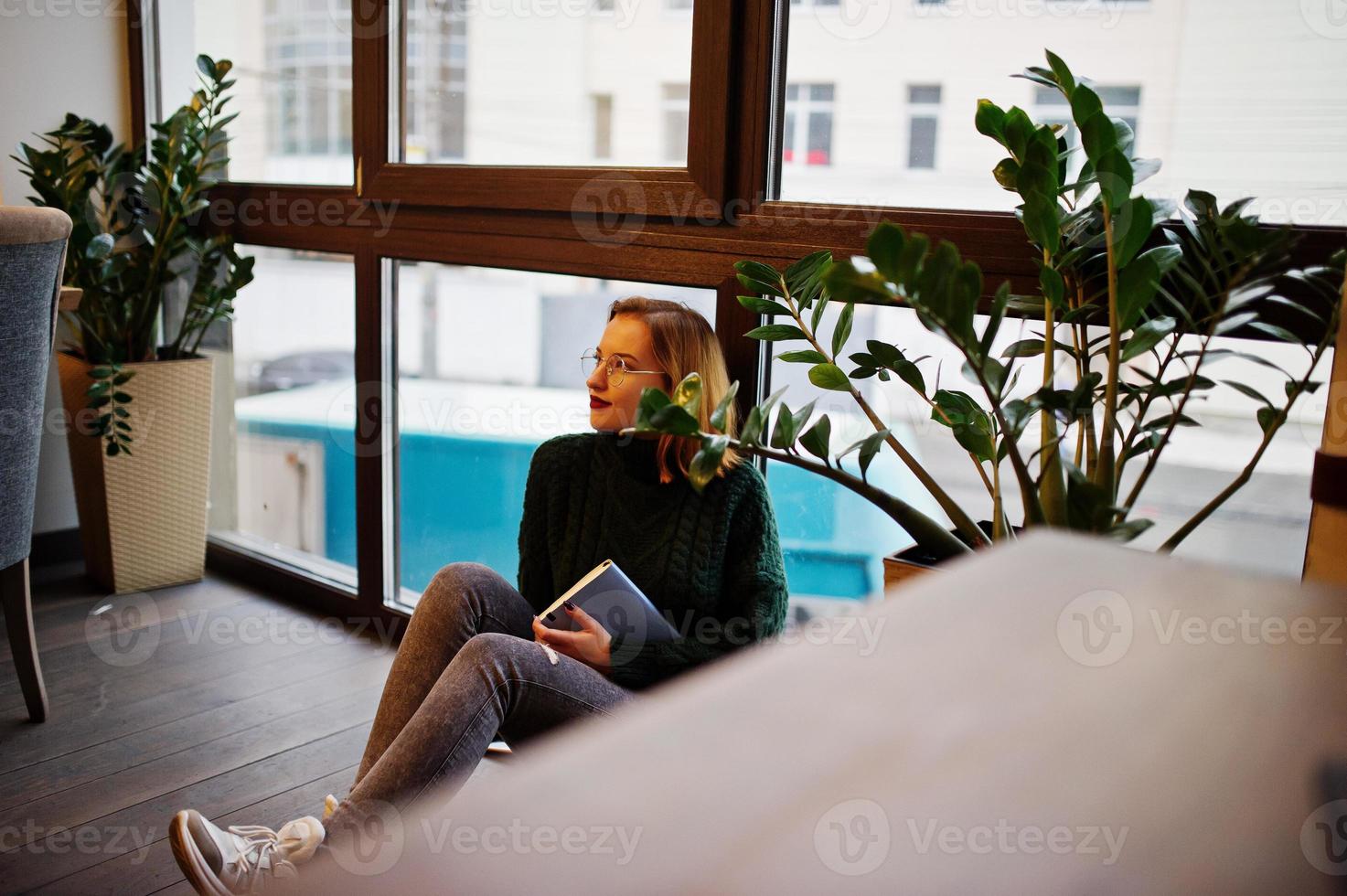 Cheerful young beautiful redhaired woman in glasses, green warm wool sweater, using her notebook, while sitting on cafe against window. She sit on floor and think about something. photo