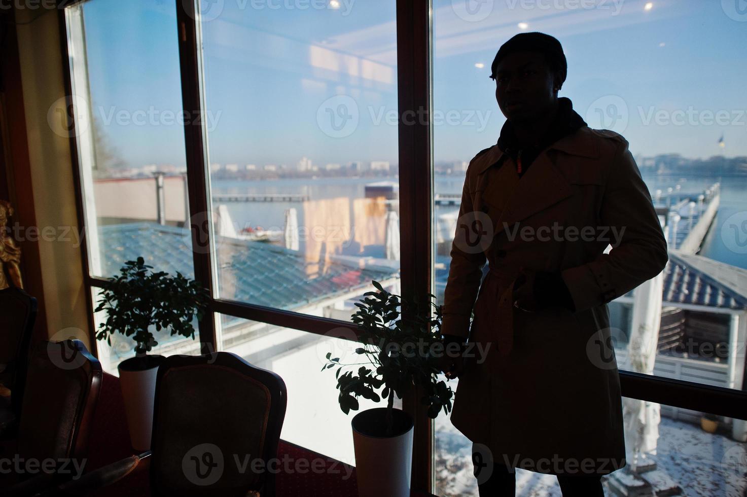 Handsome african american man posing  inside room with sunlight shadows in black hat and beige coat. photo