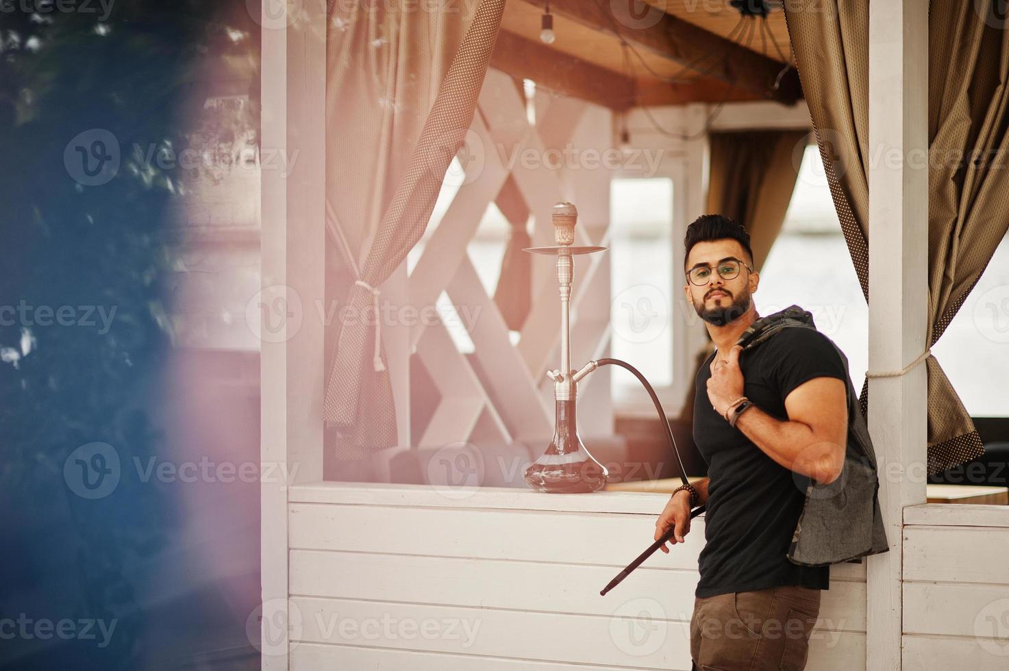 Stylish beard arabian man in glasses and black t-shirt smoking hookah outdoor. Arab model having rest. photo