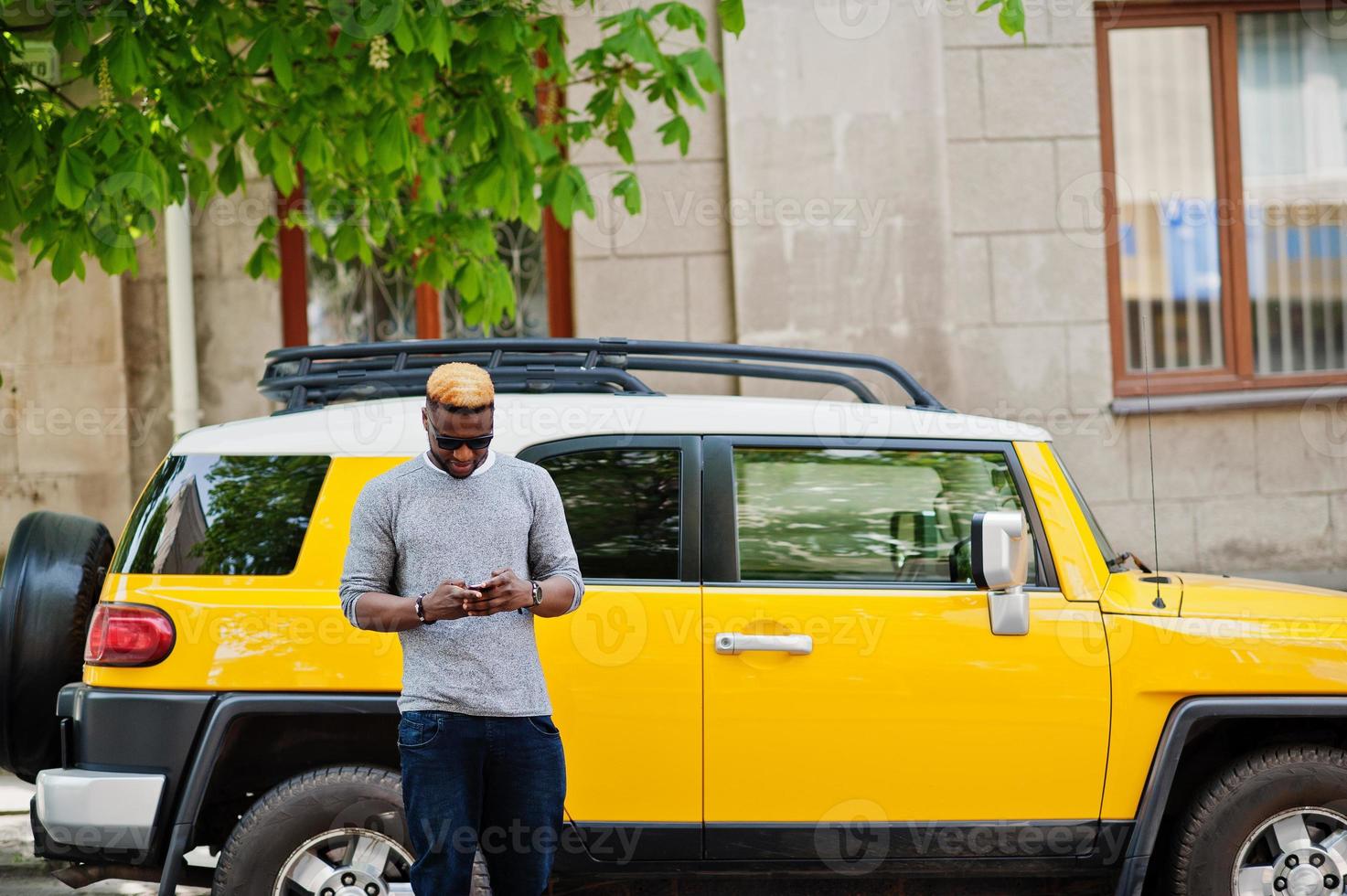 Stylish african american boy on gray sweater and black sunglasses posed on street against yellow car. Fashionable black business man. photo
