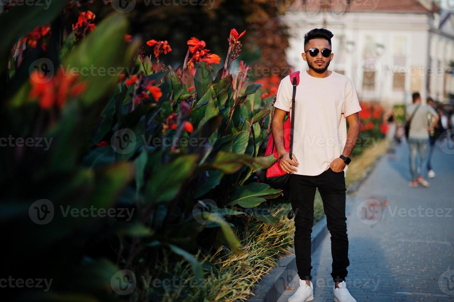 Stylish indian beard man at pink t-shirt, sunglasses and backpack. India model posed outdoor at streets of sunset city. photo