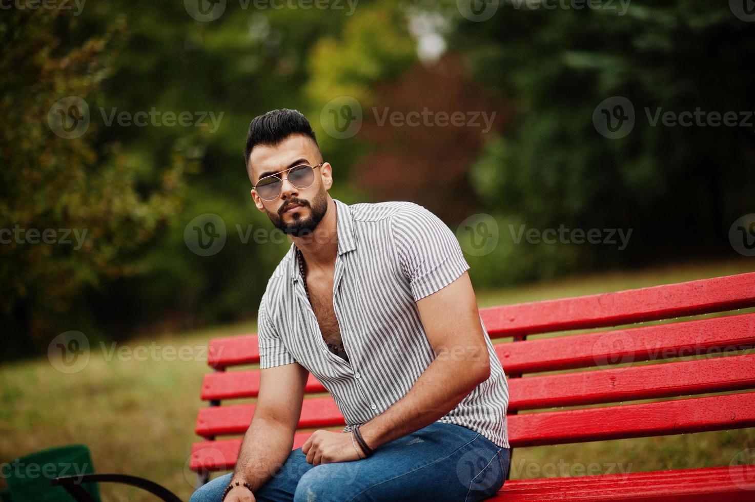 el hombre de barba árabe alto de moda usa camisa, jeans y gafas de sol sentado en un banco rojo en el parque. foto