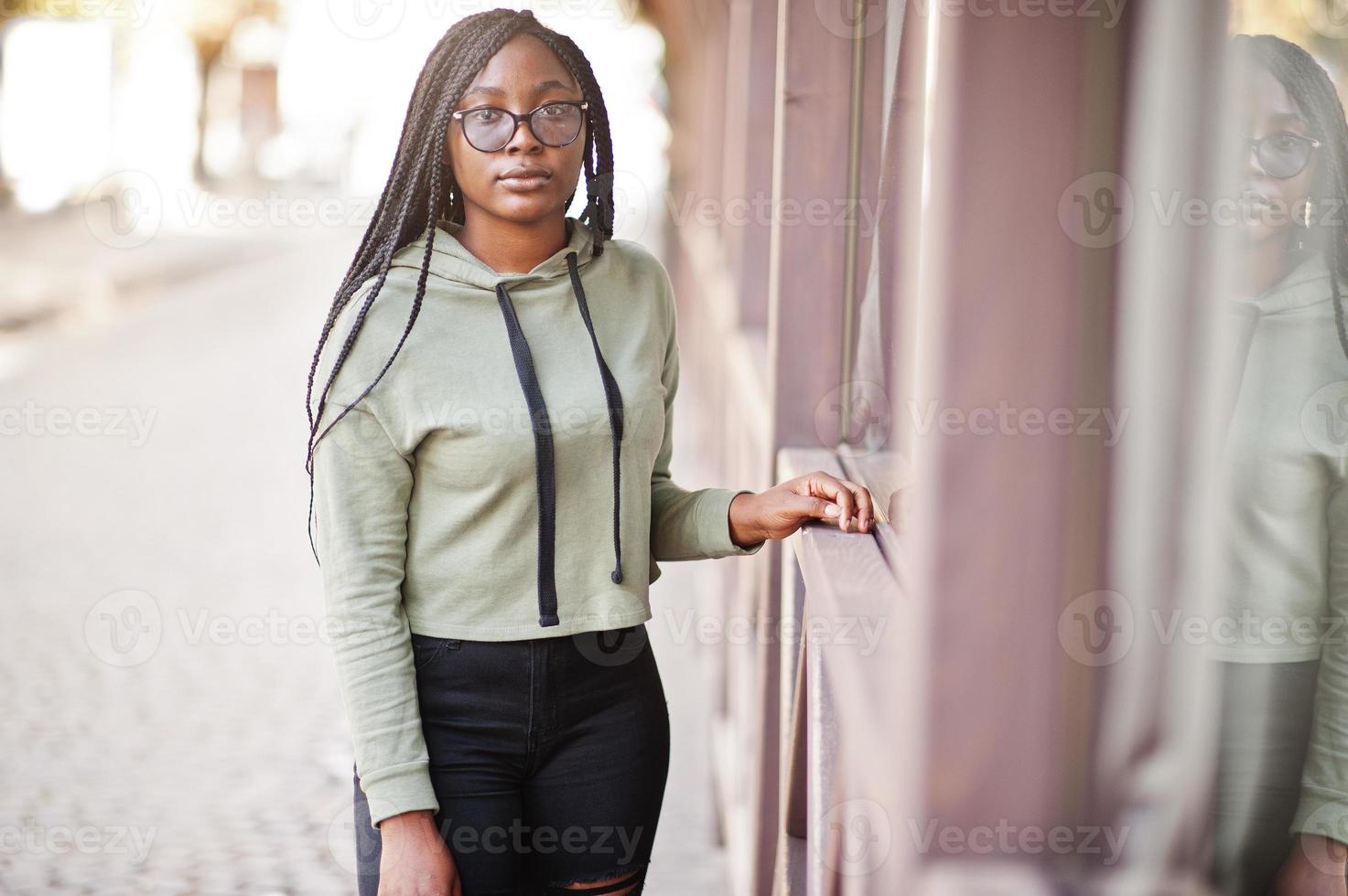 retrato de la ciudad de una mujer joven positiva de piel oscura que usa una capucha verde y anteojos. foto