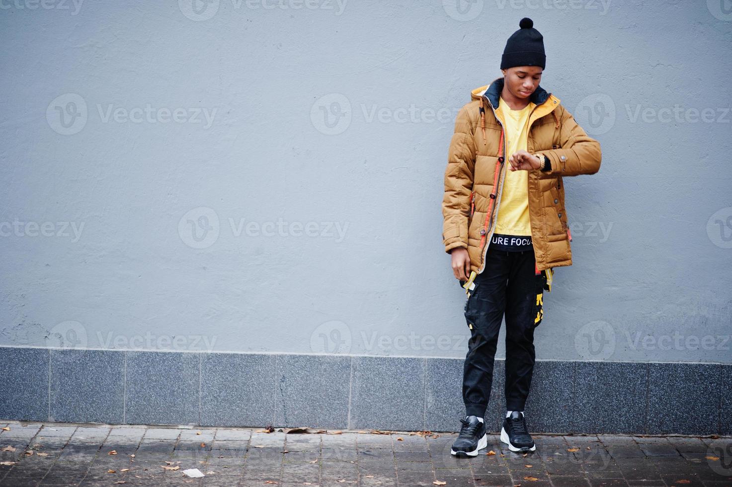 African man wear on jacket at cold weather posed outdoor against grey wall and look at his watches. photo