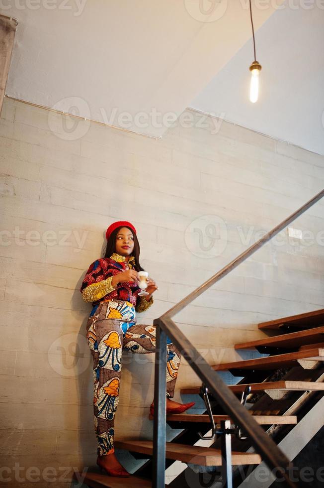 Enthusiastic african american woman in trendy coloured outfit with red beret chilling in cozy cafe, standing on stairs with cup of hot drink in hands. photo