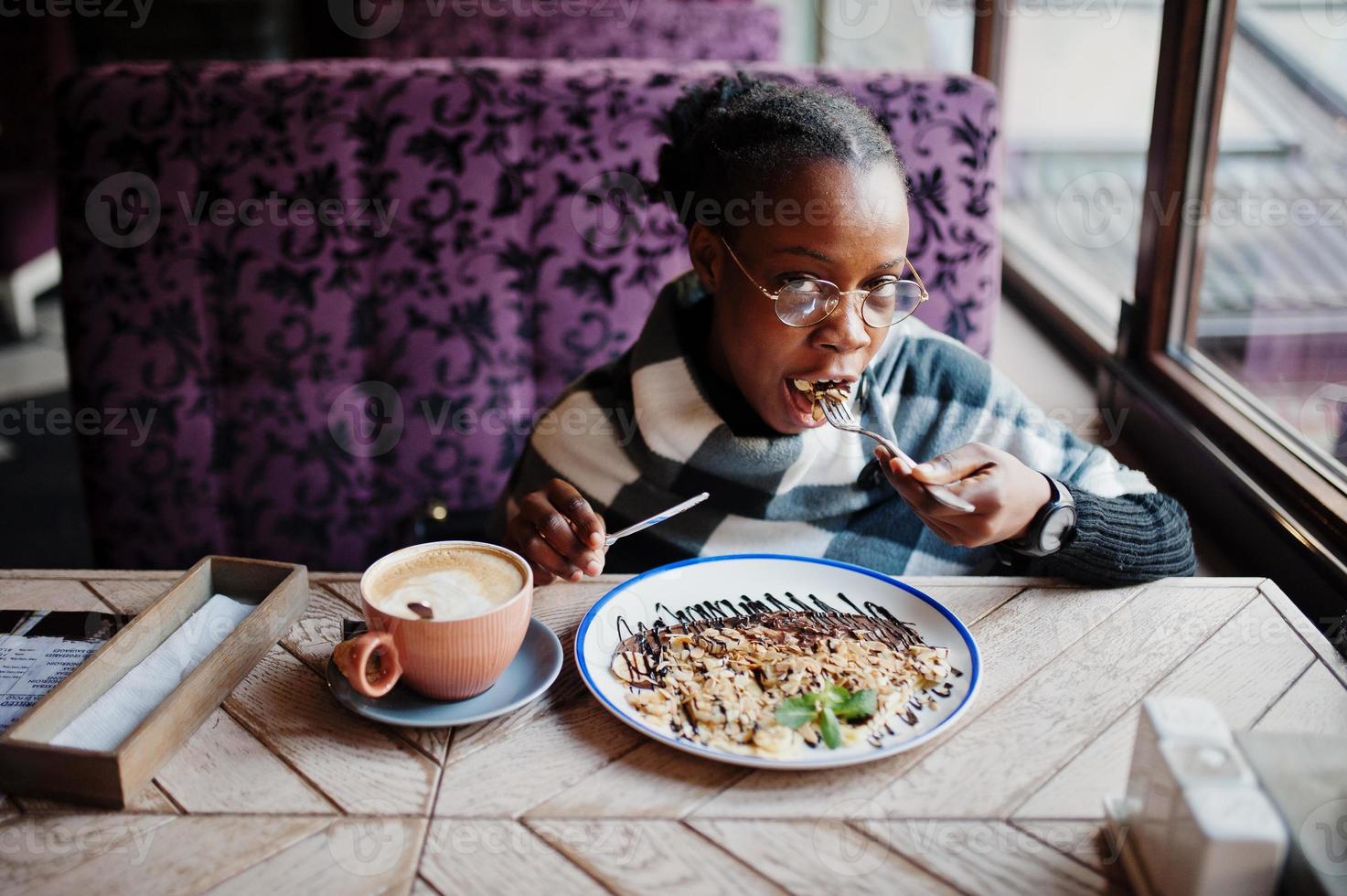 African woman in checkered cape and eyeglasses sitting at cafe and eating dessert. photo