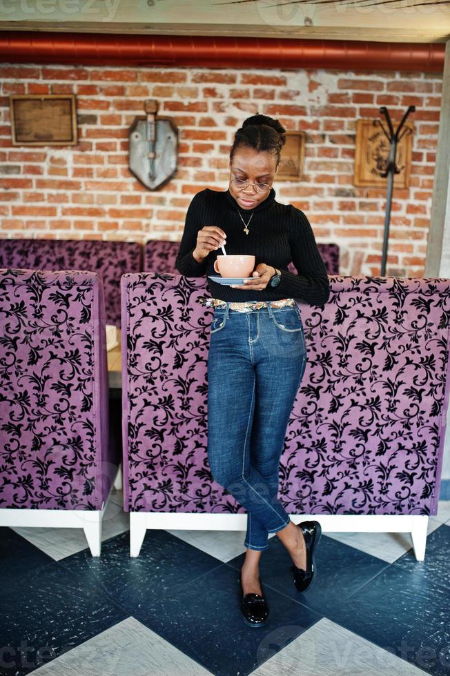 African woman in black sweater and eyeglasses posed at cafe with cup of hot drink. photo