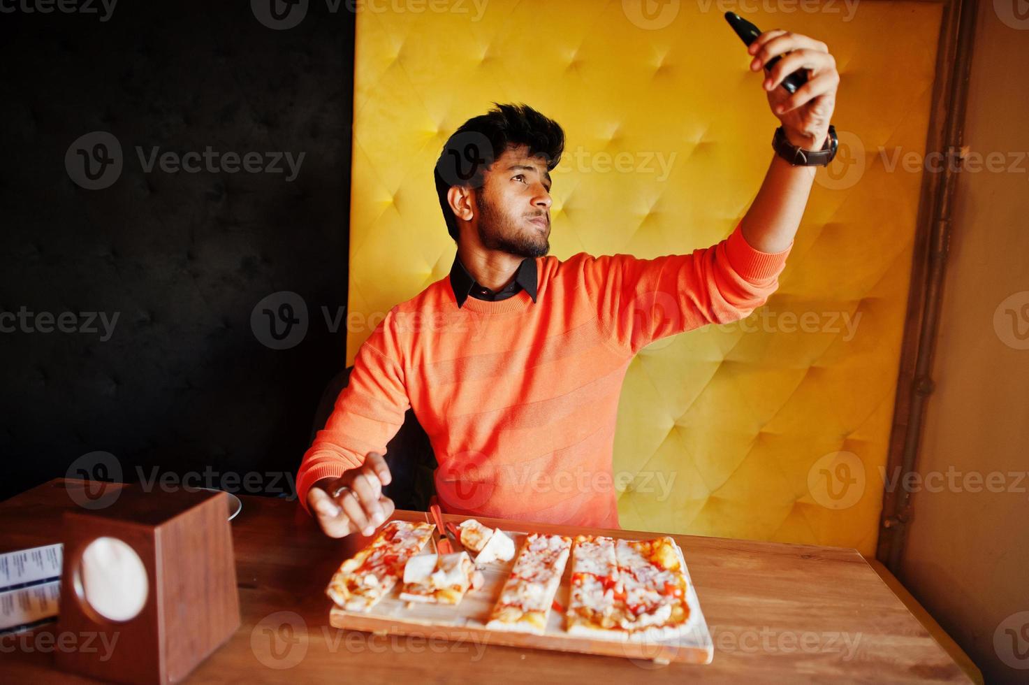 Confident young indian man in orange sweater sitting at pizzeria, eat pizza and makes photo on his phone.