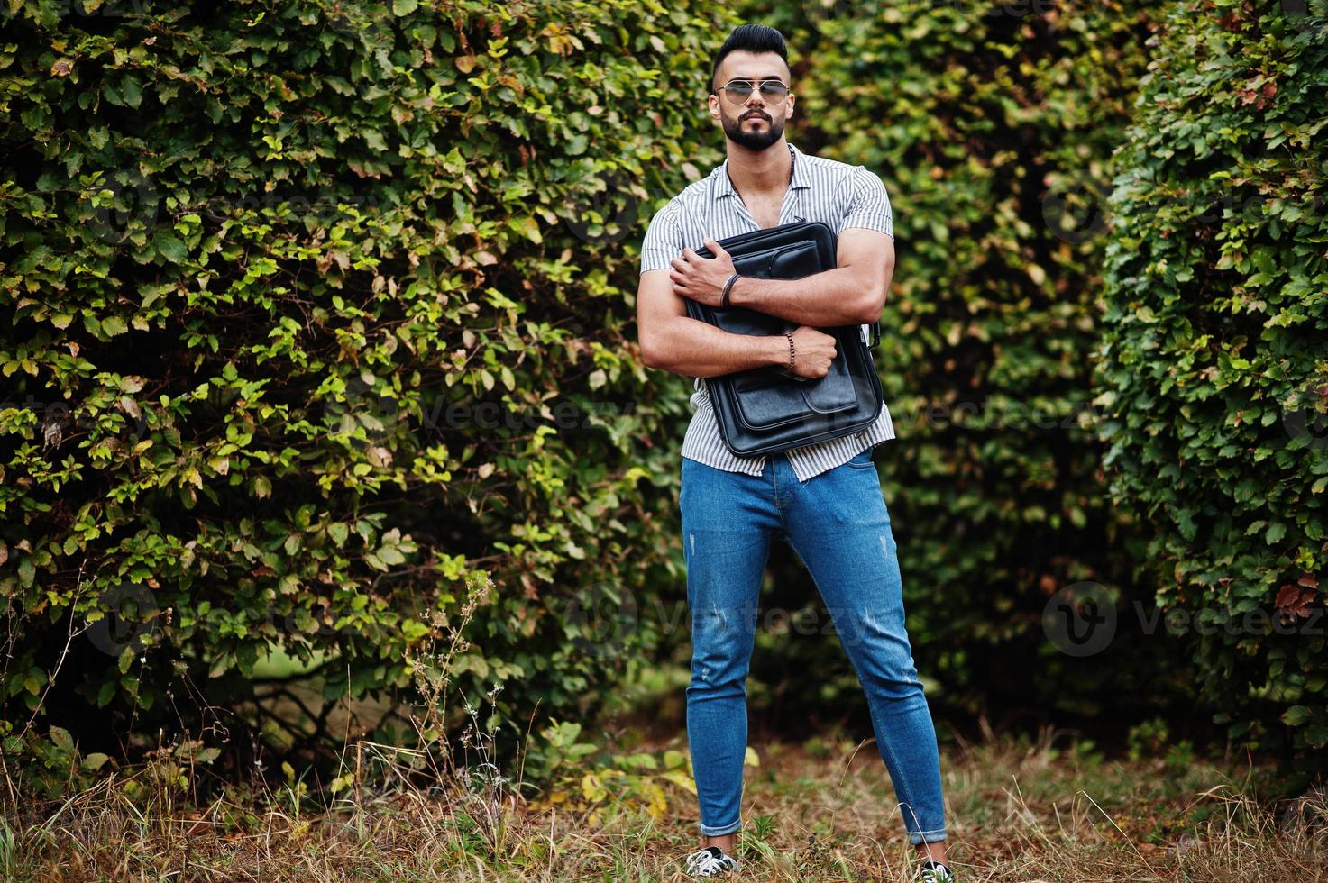 el hombre de barba árabe alto de moda usa camisa, jeans y gafas de sol posados en el parque y sostiene la caja de la bolsa. foto