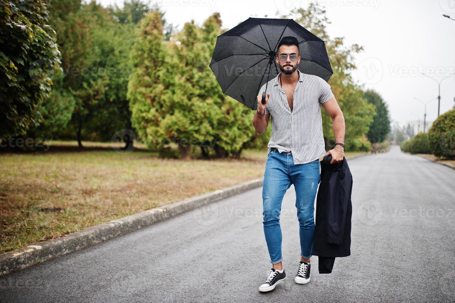 Fashionable tall arab beard man wear on shirt, jeans and sunglasses walking at park with umbrella and coat at hand. photo