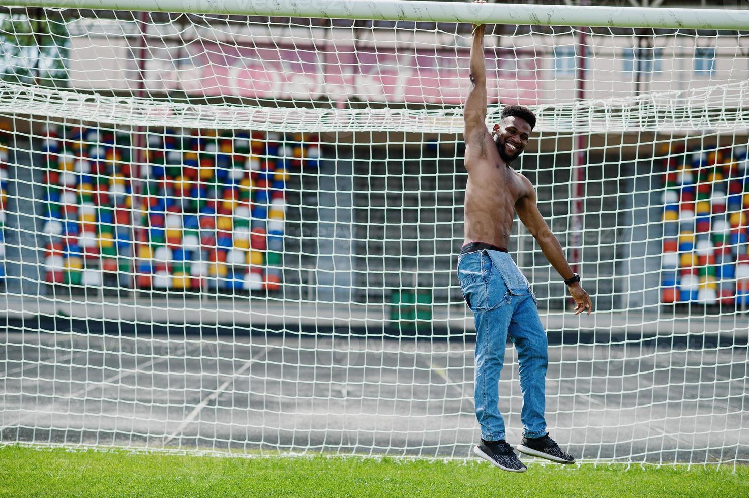 Handsome sexy african american muscular bare torso man at jeans overalls posed at green grass agasinst football gates of stadium field. Fashionable black man portrait. photo