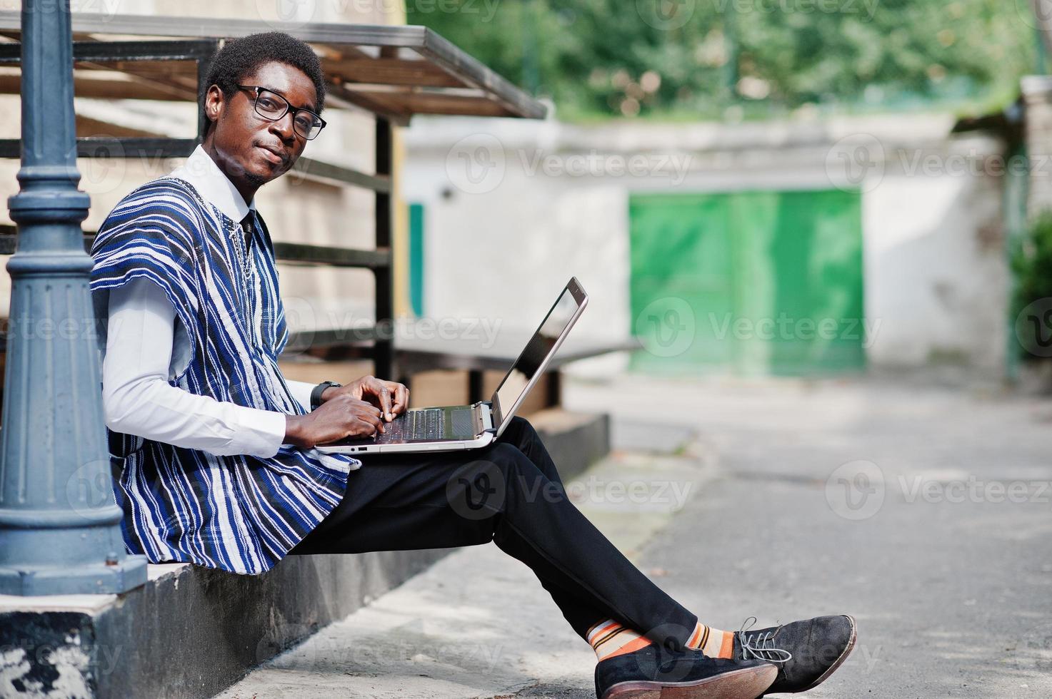 hombre africano con ropa tradicional y gafas con laptop trabajando al aire libre. foto