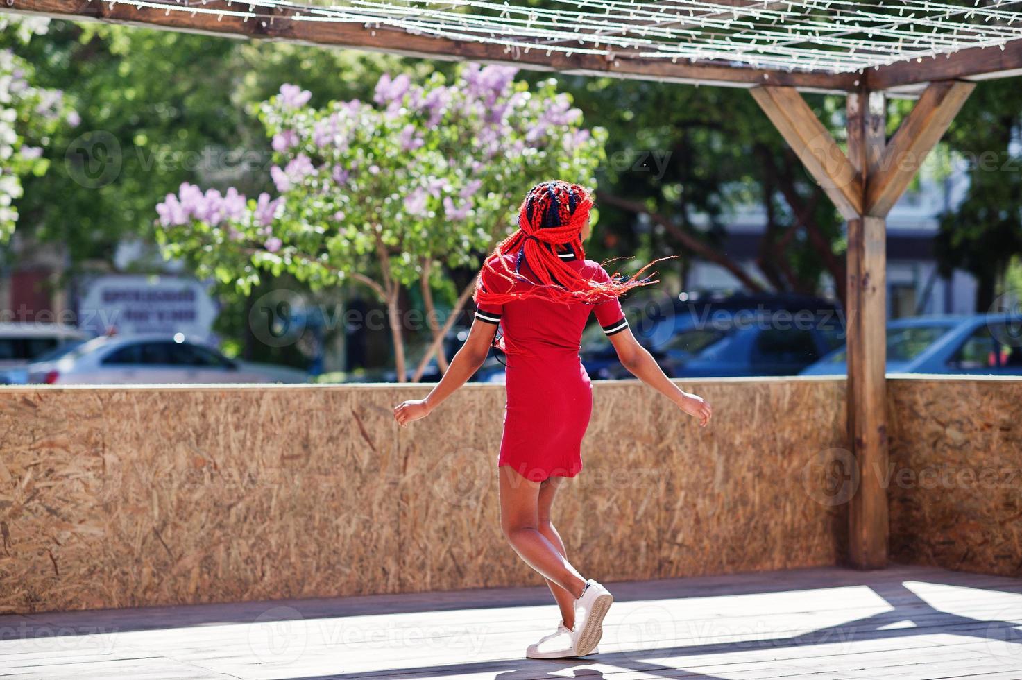 Cute and slim african american girl in red dress with dreadlocks in motion having fun on street. Stylish black model. photo