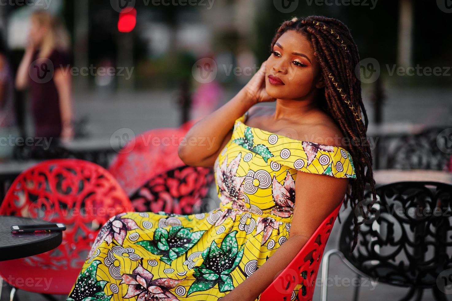 linda chica afroamericana de pequeña altura con rastas, vestida con un vestido amarillo de color, sentada en un café al aire libre en una silla roja. foto