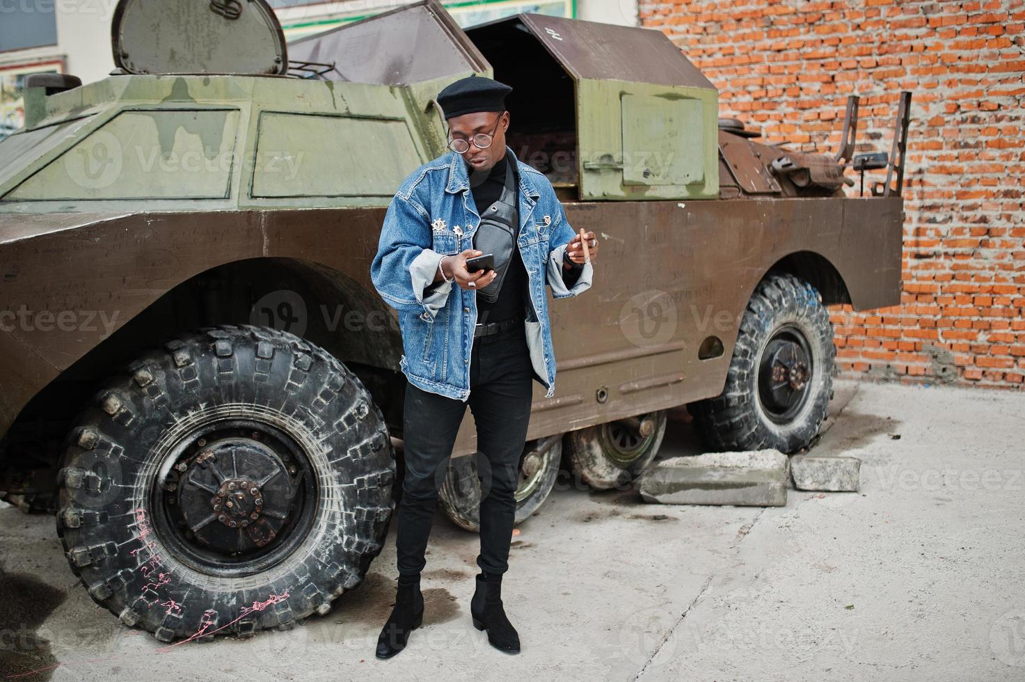 African american man in jeans jacket, beret and eyeglasses, smoking cigar and posed against btr military armored vehicle, with mobile phone at hand. photo