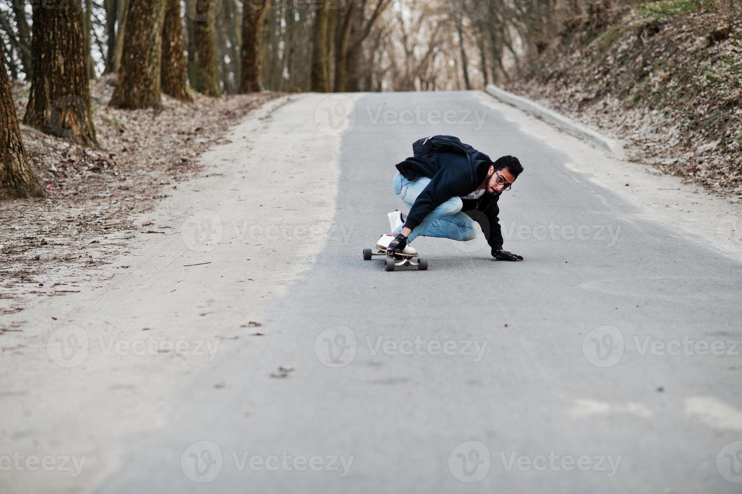 Street style arab man in eyeglasses with longboard longboarding down the road. photo