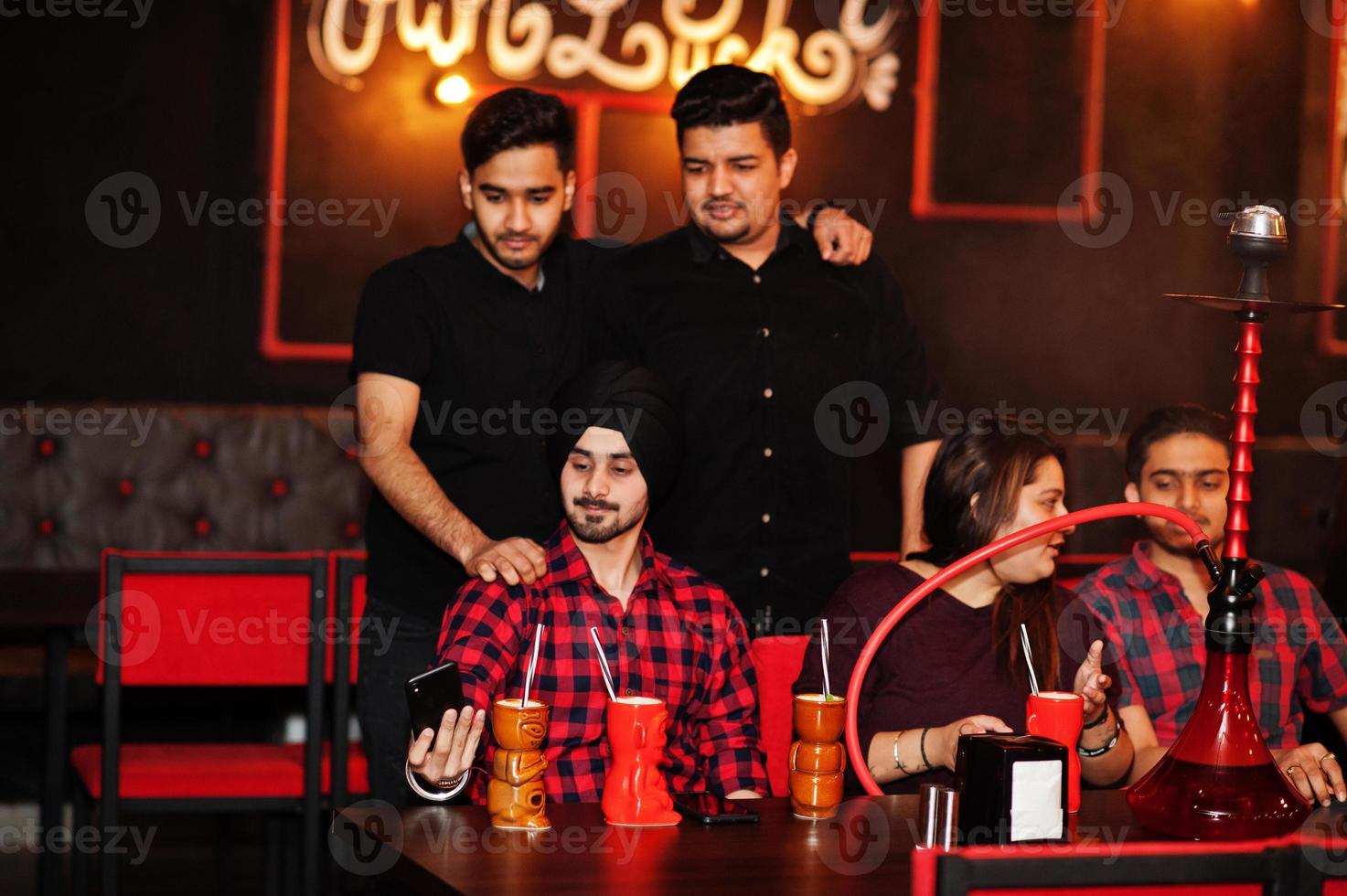 Group of indian friends having fun and rest at night club, drinking cocktails and smoke hookah, looking at mobile phones. photo