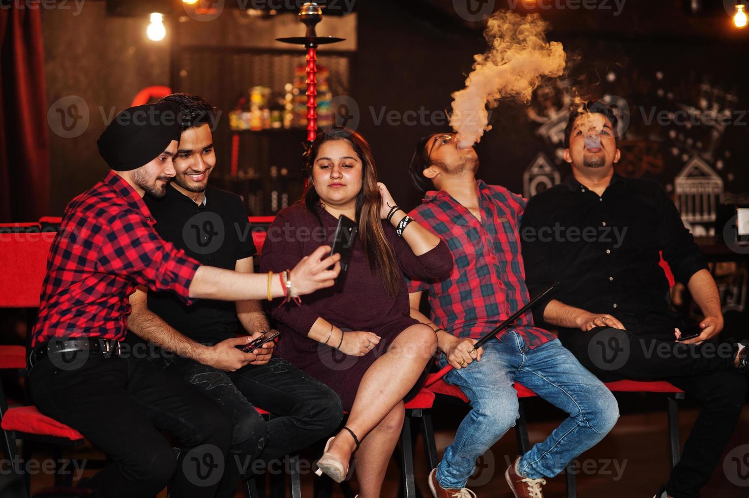 Group of indian friends sitting at lounge bar, having fun and rest, smoking hookah and looking on mobile phones to make selfie. photo