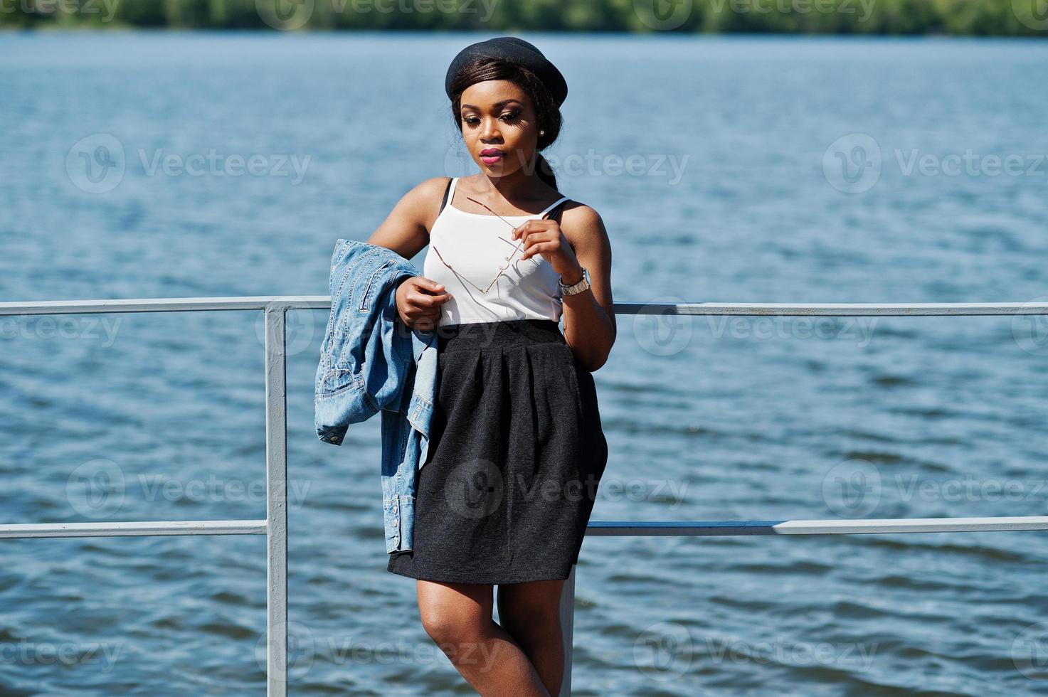 elegante modelo afroamericano con sombrero de anteojos, chaqueta de jeans y  falda negra posada al aire libre en el muelle contra el lago. 10431205 Foto  de stock en Vecteezy