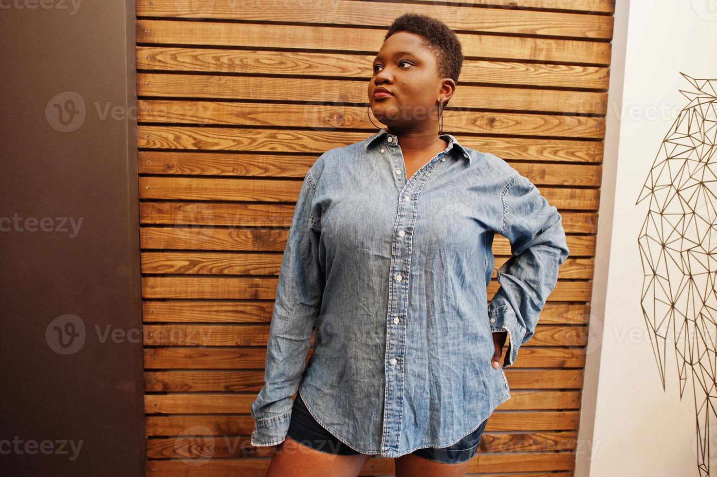Casual african woman in jeans shirt and shots clothes posing against wooden wall. photo