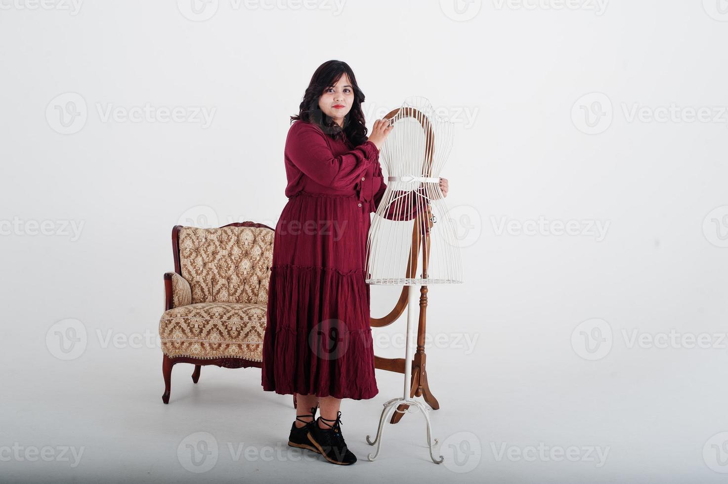 Attractive south asian woman in deep red gown dress posed at studio on white background against mirror and chair. photo