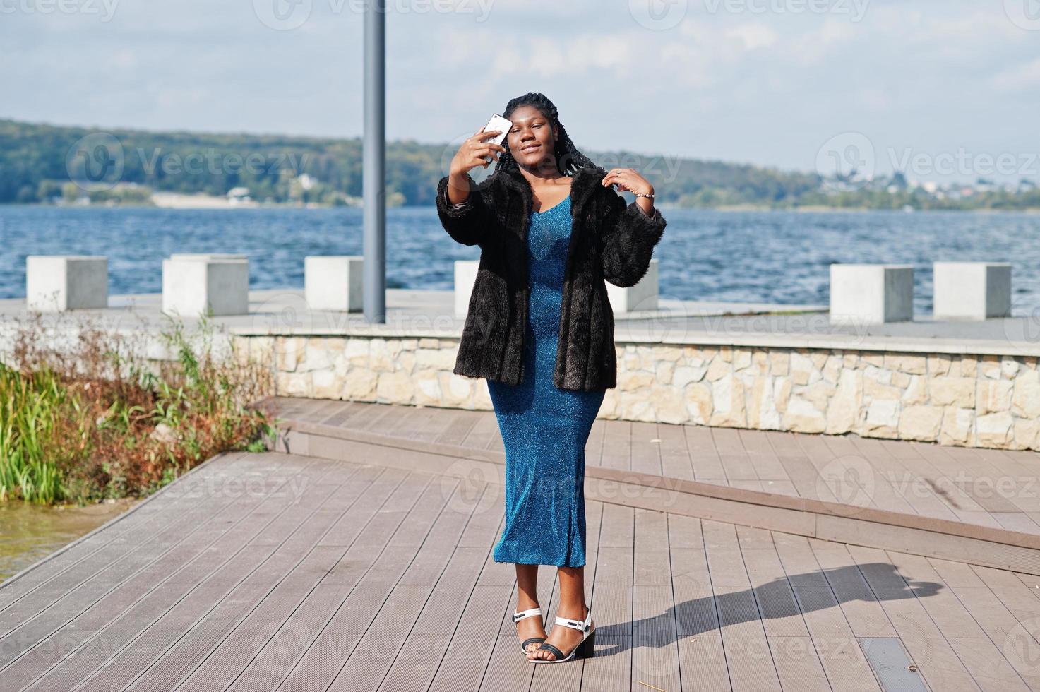 African american dark skinned plus size model posed in a blue shiny dress and black fur coat against sea side making selfie on telephone. photo