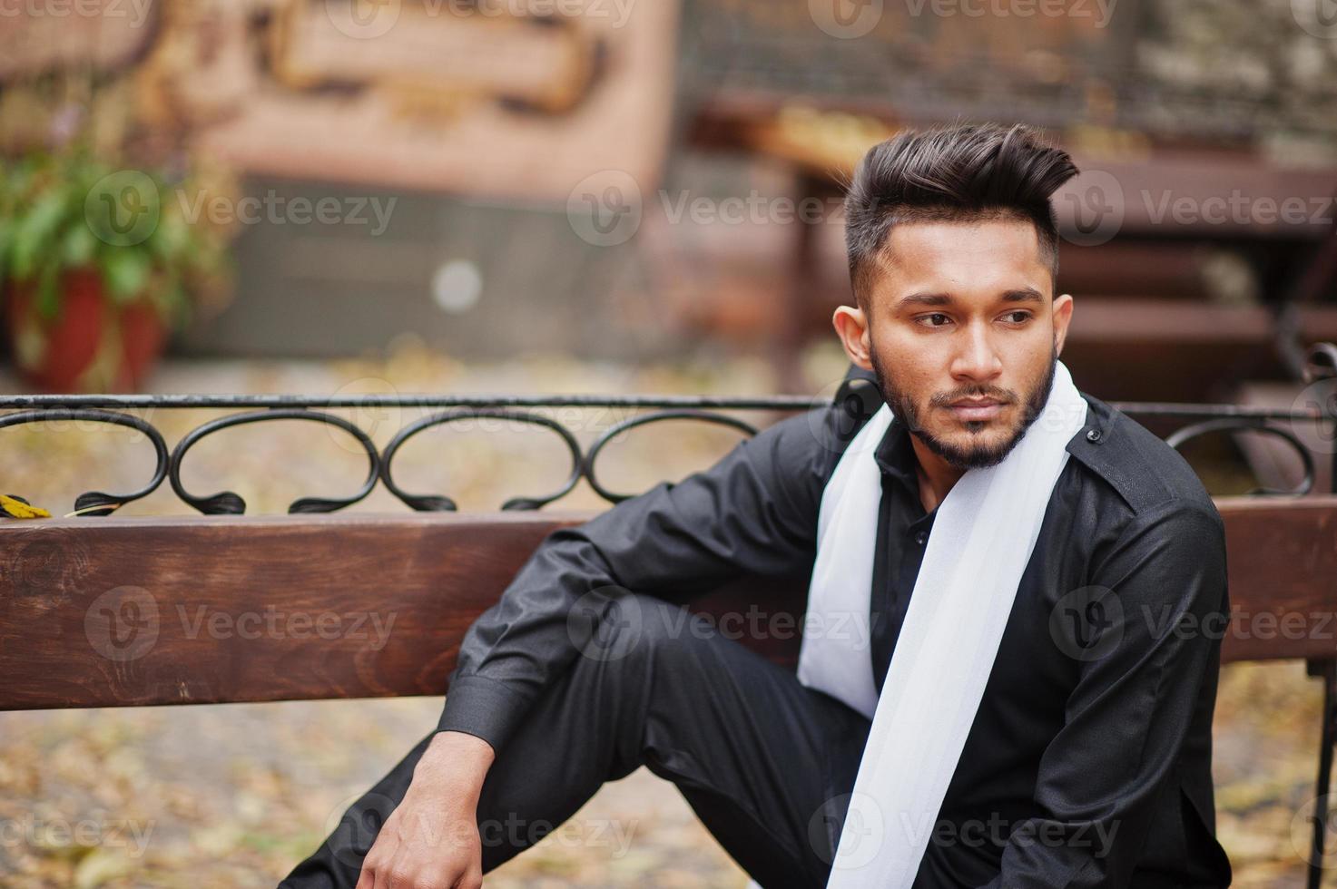 Indian stylish man in black traditional clothes with white scarf posed outdoor sitting on bench. photo