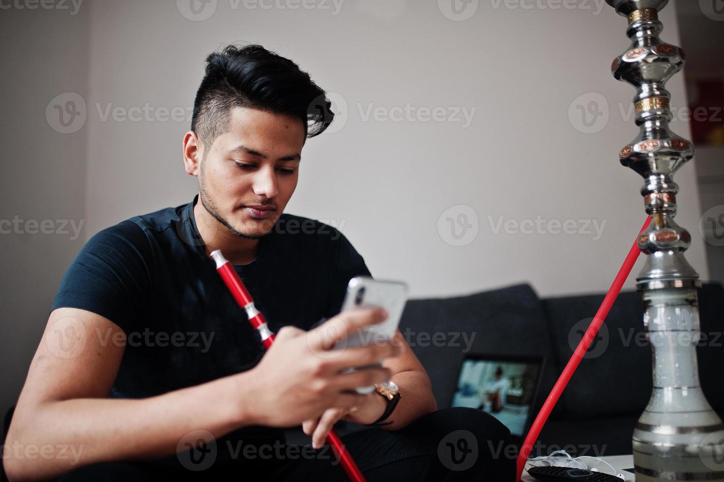 Handsome and fashionable indian man in black sitting at room, smoking hookah and looking on mobile phone. photo