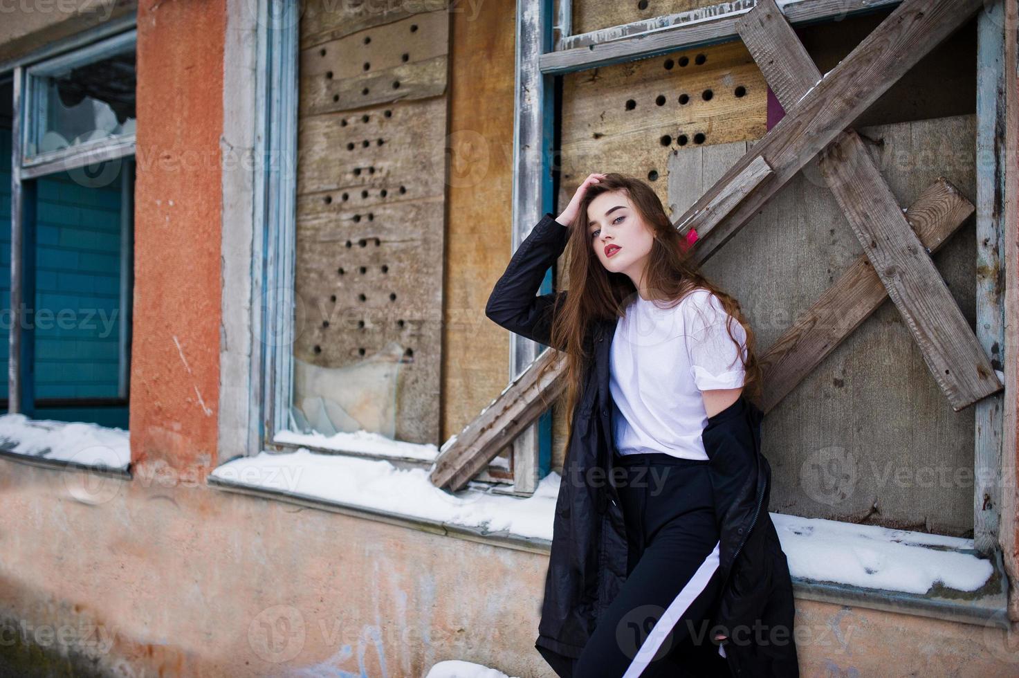 Fashionable long legs brunette model in long black cloak posed outdoor at winter day against old grunge wall with broken windows. photo