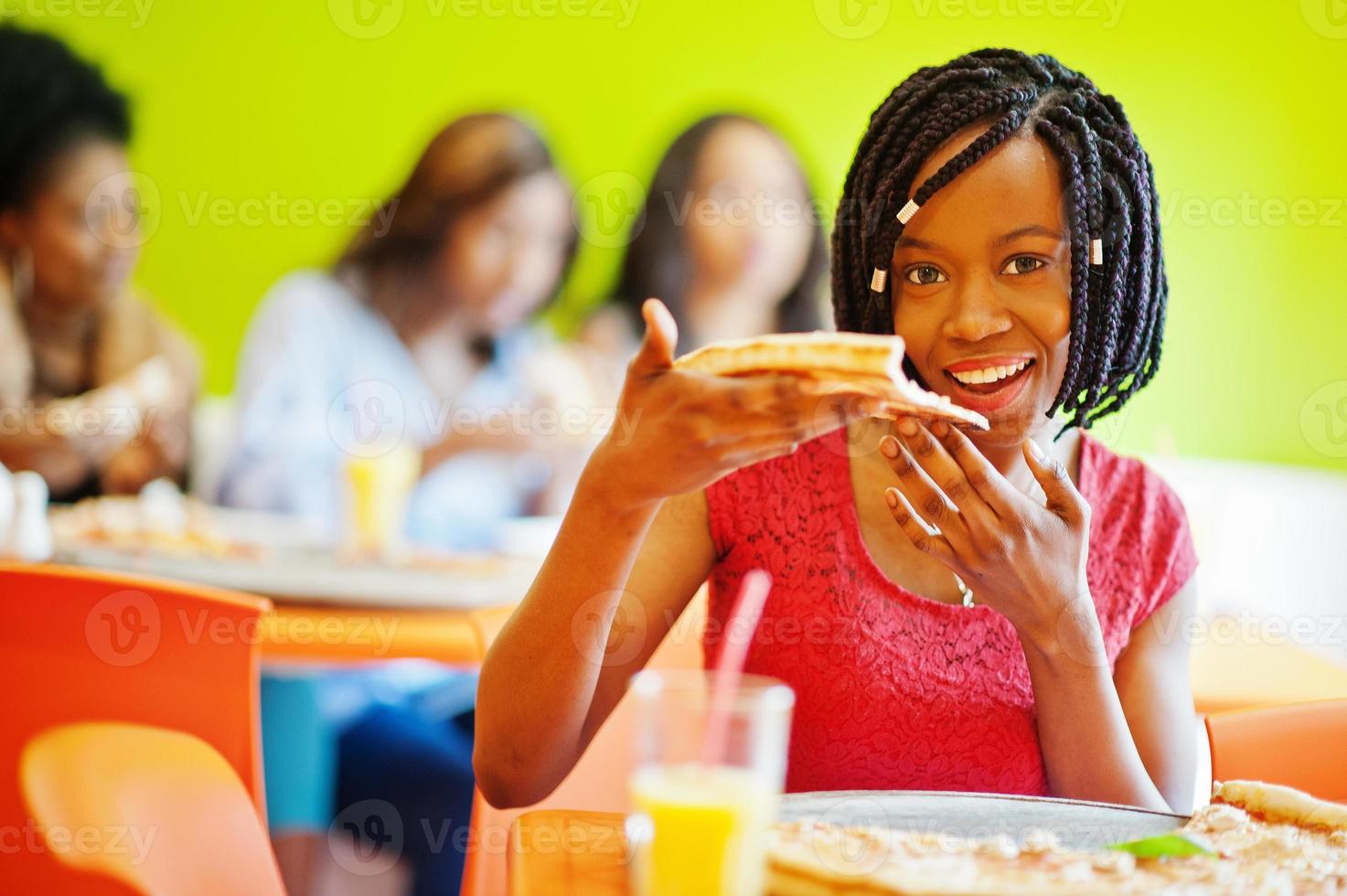 African woman with pizza sitting at restaurant against dark skinned girls. photo