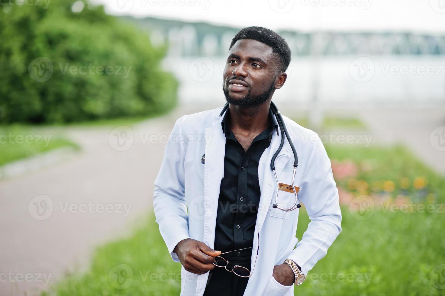 joven médico afroamericano de bata blanca con un estetoscopio posado al aire libre. foto