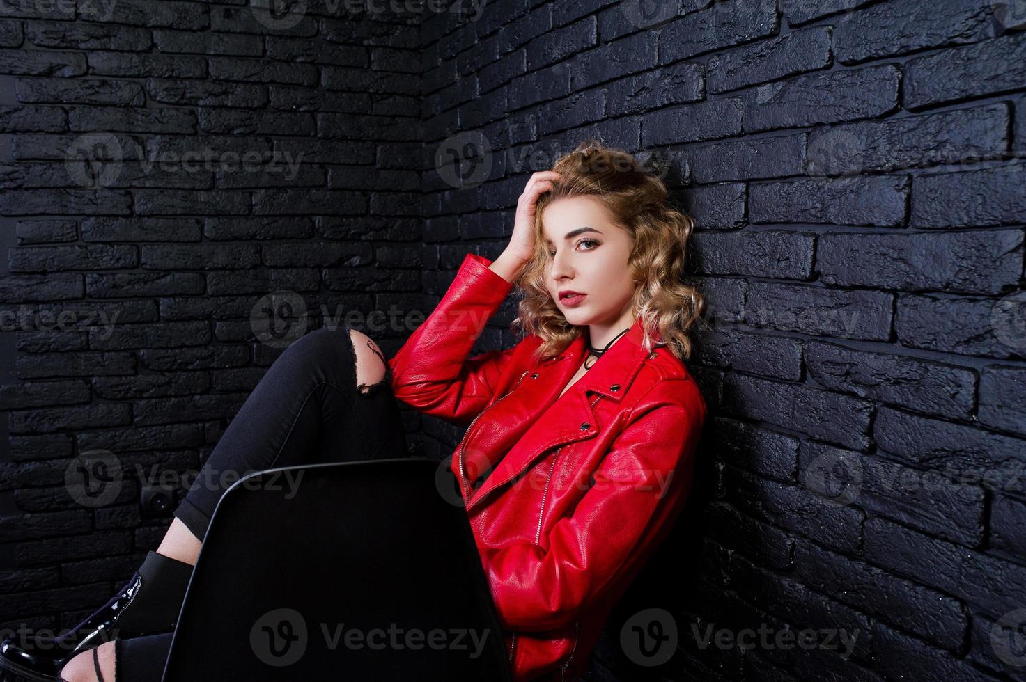 Studio portrait of blonde girl in red leather jacket posed on chair against brick wall. photo