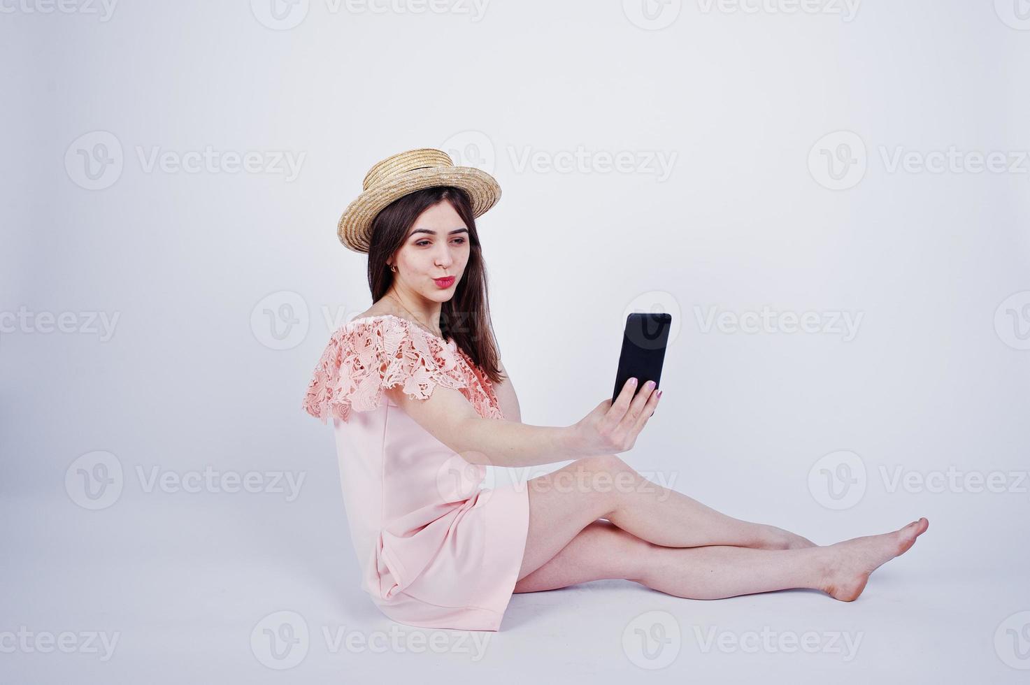 Portrait of a fashionable woman in pink dress and hat sitting on the floor and taking selfie in the studio. photo