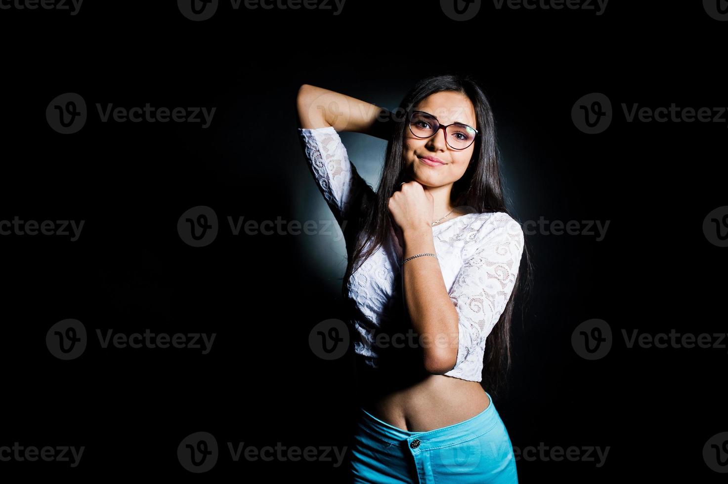 Portrait of an attractive young woman in white top and blue pants posing with her glasses in the dark. photo
