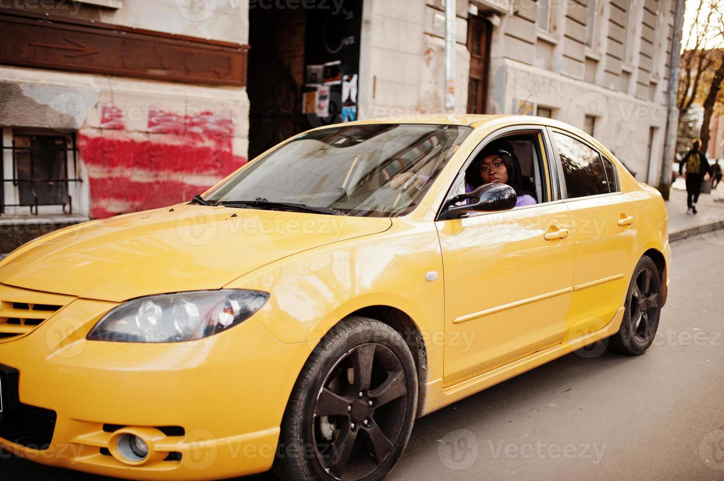 African american woman at violet dress and cap posed at yellow car. photo