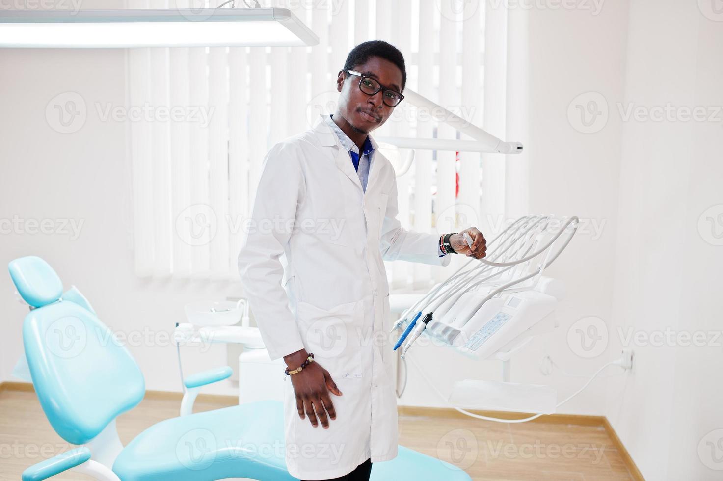 médico afroamericano con anteojos parado cerca de la silla del dentista en la clínica dental. foto
