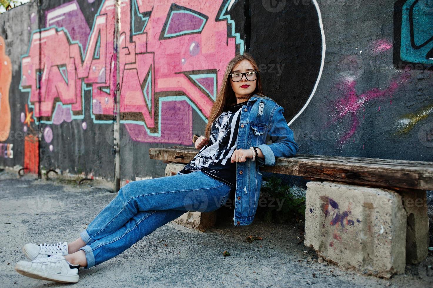 chica hipster casual con estilo en ropa de jeans y gafas contra una gran pared de graffiti. foto